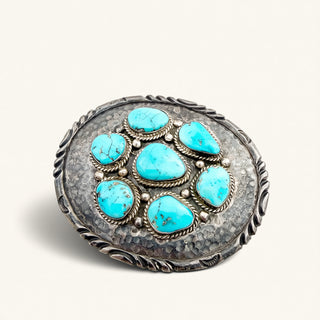 A circular silver-toned jewelry piece featuring seven turquoise stones arranged in a cluster, photographed on a clean off-white background.