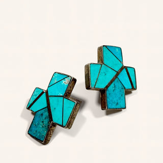 A pair of geometric cross-shaped cufflinks featuring inlay turquoise stones in a mosaic pattern, set in dark metal on a plain off-white background.