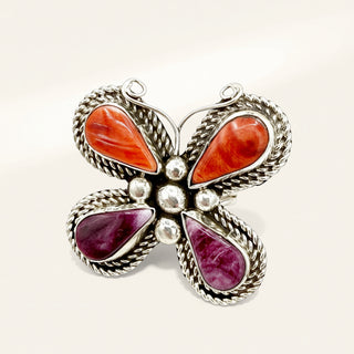 A butterfly-shaped silver ring featuring four teardrop-shaped gemstones in orange and purple, with a textured silver border and small silver beads at the center, set against a plain light background.